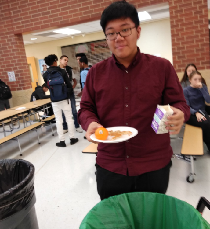 Teenaged boy carrying empty milk carton and plate, preparing to deposit materials into waste sorting bins. Tables, other students, and other features of the school cafeteria are in background.